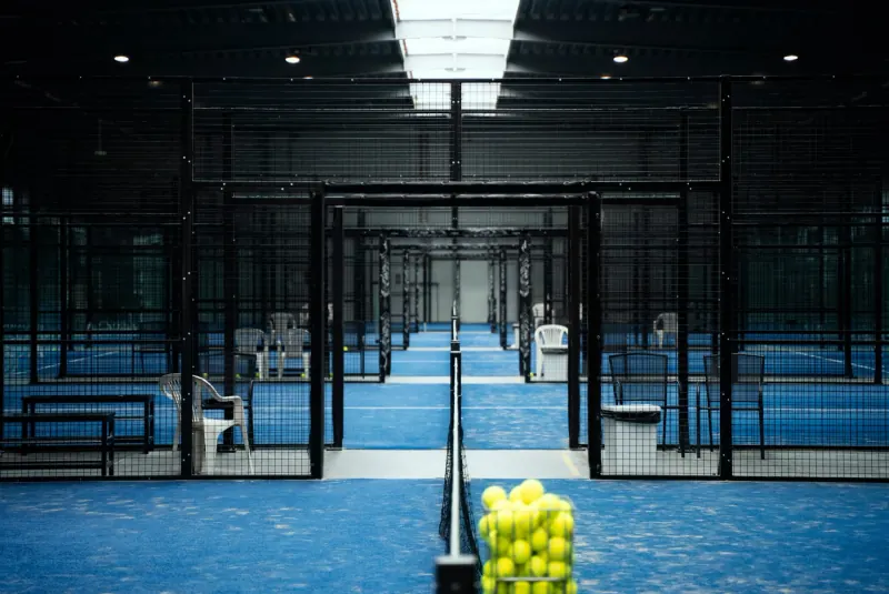A row of indoor padel courts seen through their steel mesh frames, with a basket of balls by the net