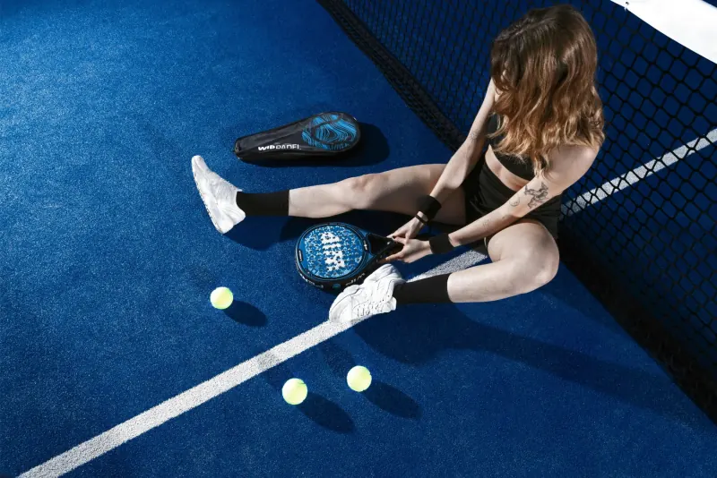 A padel player sitting by the net on a blue court with her racket and balls