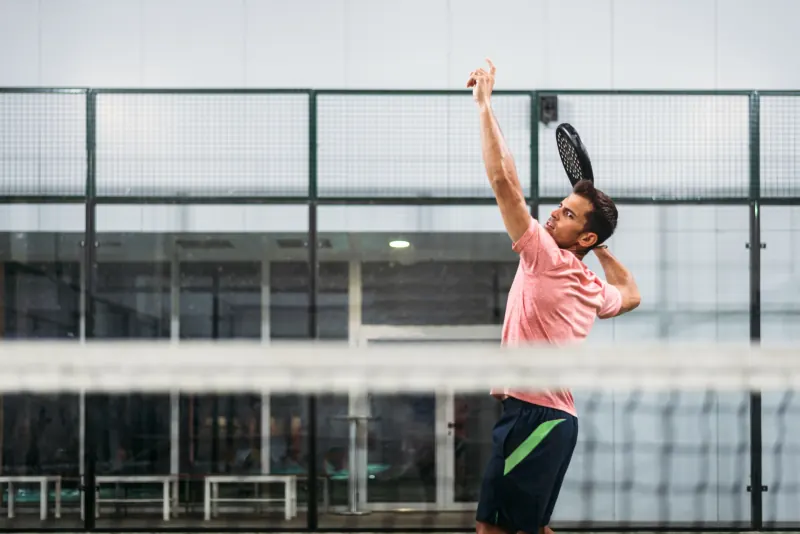 A padel player in a pink shirt reaching up to serve on an indoor court